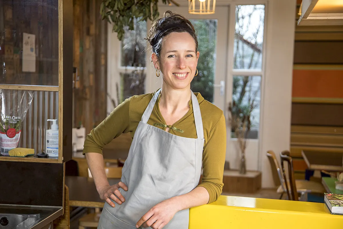 Een vrouw met donker achterovergebonden haar, gekleed in een groen shirt met lange mouwen en een grijs schort, staat met één hand op haar heup en de andere op een geel aanrecht. Achter haar staat een houten plank met diverse spullen en een eethoek met stoelen en een raam met een buitentafereel. Zij lacht.