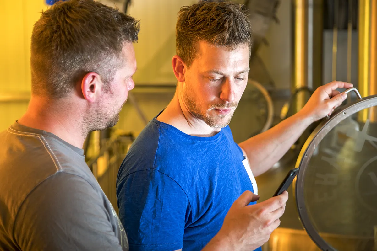 Twee mannen in een brouwerij raken in gesprek. De man rechts, gekleed in een blauw shirt, houdt een smartphone vast en lijkt gefocust op het scherm. De man links, gekleed in een grijs overhemd, kijkt hem aandachtig aan. Op de achtergrond is industriële brouwapparatuur zichtbaar.