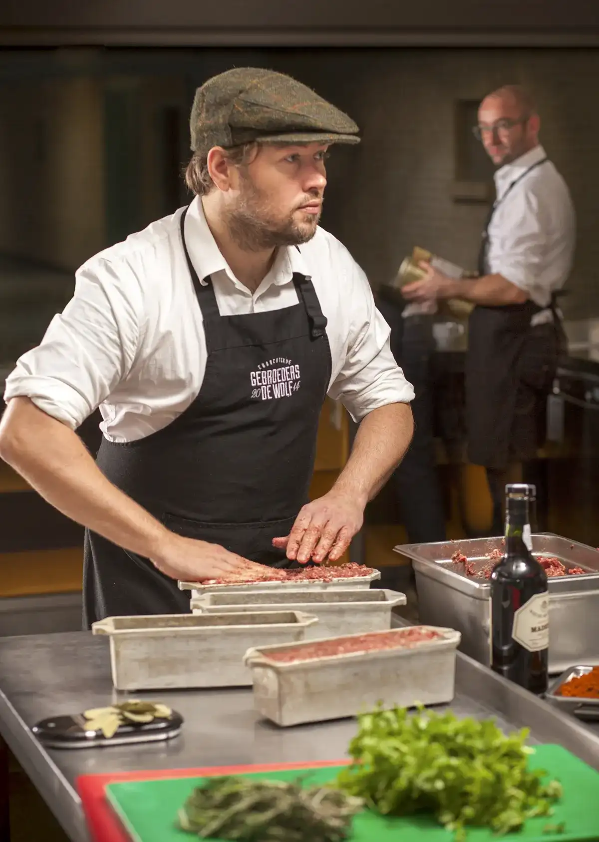 Een man met een platte pet en een zwart schort met de tekst "Georges Descrières" drukt gemalen vlees in rechthoekige metalen mallen op een roestvrijstalen aanrecht. Een andere man op de achtergrond maakt het eten klaar. Op het aanrecht staan diverse kookingrediënten, een wijnfles, kruiden en een snijplank.