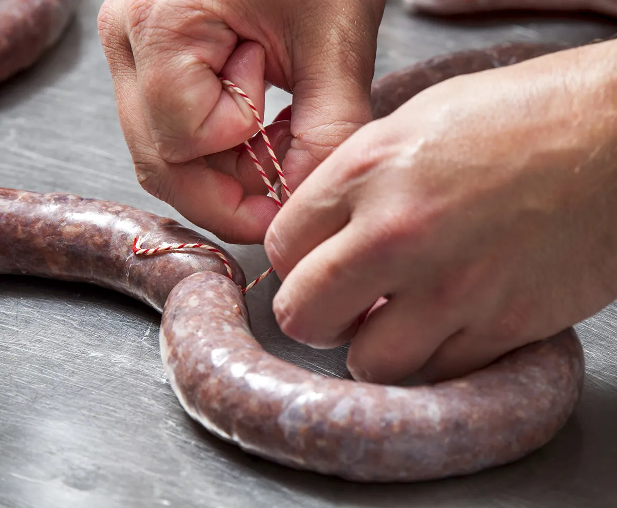 Close-up van handen die een dikke worst binden met rood en wit touw op een roestvrijstalen oppervlak. De worst is gebogen en gedeeltelijk afgesneden terwijl het touw in een lus wordt gelegd en wordt vastgezet. De persoon is bezig met het bereiden of afwerken van de zelfgemaakte worst.