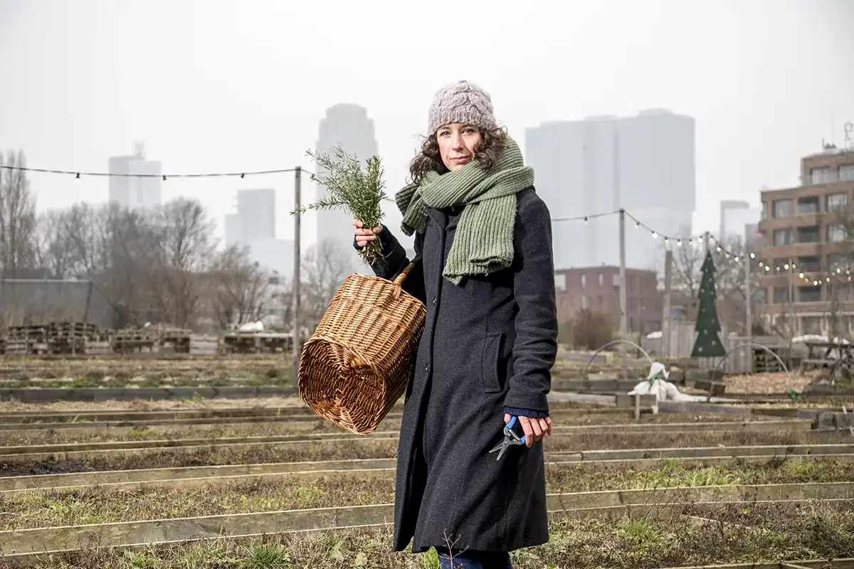 Een vrouw in een donkere jas en groene sjaal, met een rieten mand en een kleine tak in haar handen, staat in een stadstuin met verhoogde plantbedden. Ze draagt een grijze gebreide muts en kijkt naar de camera. De achtergrond omvat bladloze bomen, stadsgebouwen en lichtslingers. Bewolkte hemel boven.