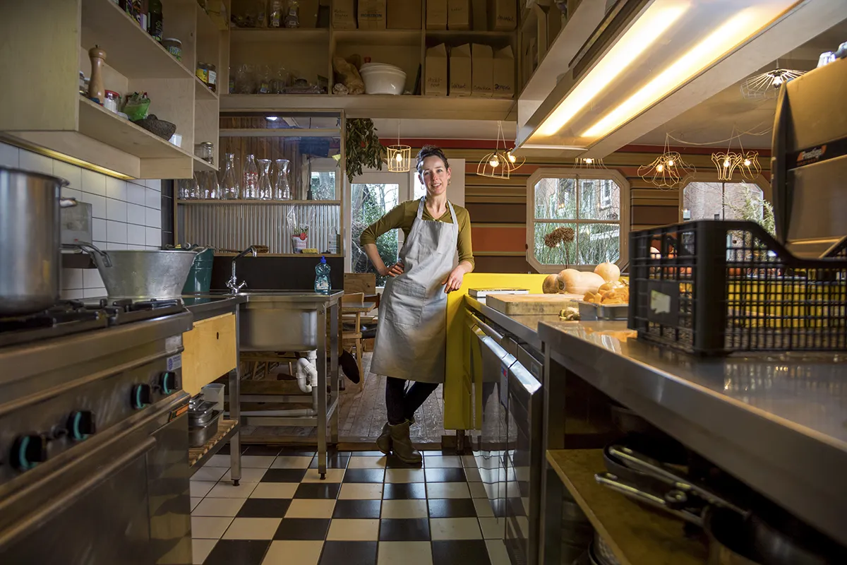 Een vrouw met een grijs schort staat zelfverzekerd in een professionele keuken. Ze laat haar arm op een metalen aanrecht rusten, omringd door kookgerei en keukengerei. De keuken heeft een geblokte zwart-witte vloer en op de achtergrond zijn houten planken beladen met potten en ingrediënten zichtbaar.