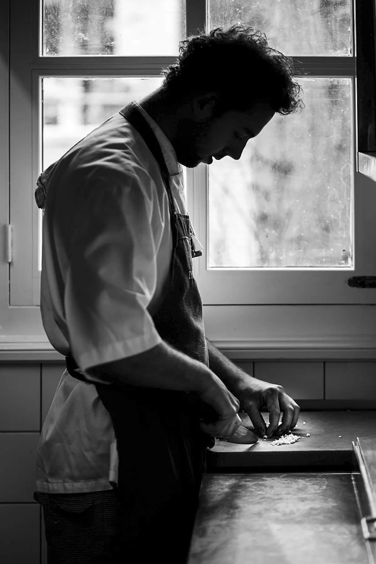 Zwart-wit beeld van een chef-kok die in een keuken staat en groenten hakt op een snijplank. De chef-kok draagt een wit overhemd en een donker schort, gericht op zijn taak. Het zonlicht stroomt door het raam achter hem, waardoor schaduwen ontstaan en een serene sfeer ontstaat.