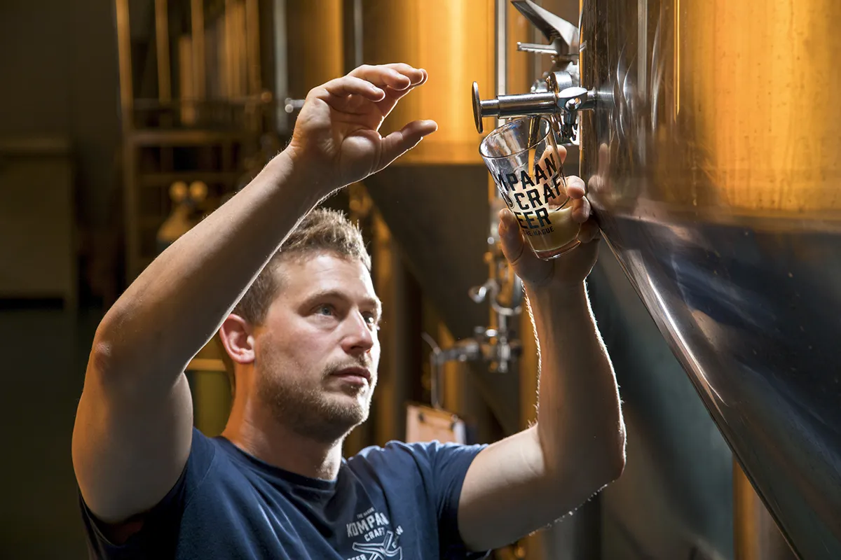Een man, gekleed in een donker overhemd, schenkt bier uit een grote roestvrijstalen tank in een helder glas met de tekst 'BREW BARIAN CRAFT BEER'. Hij lijkt gefocust op het proces, dat plaatsvindt in een industrieel ogende brouwerijomgeving met extra brouwapparatuur zichtbaar op de achtergrond.