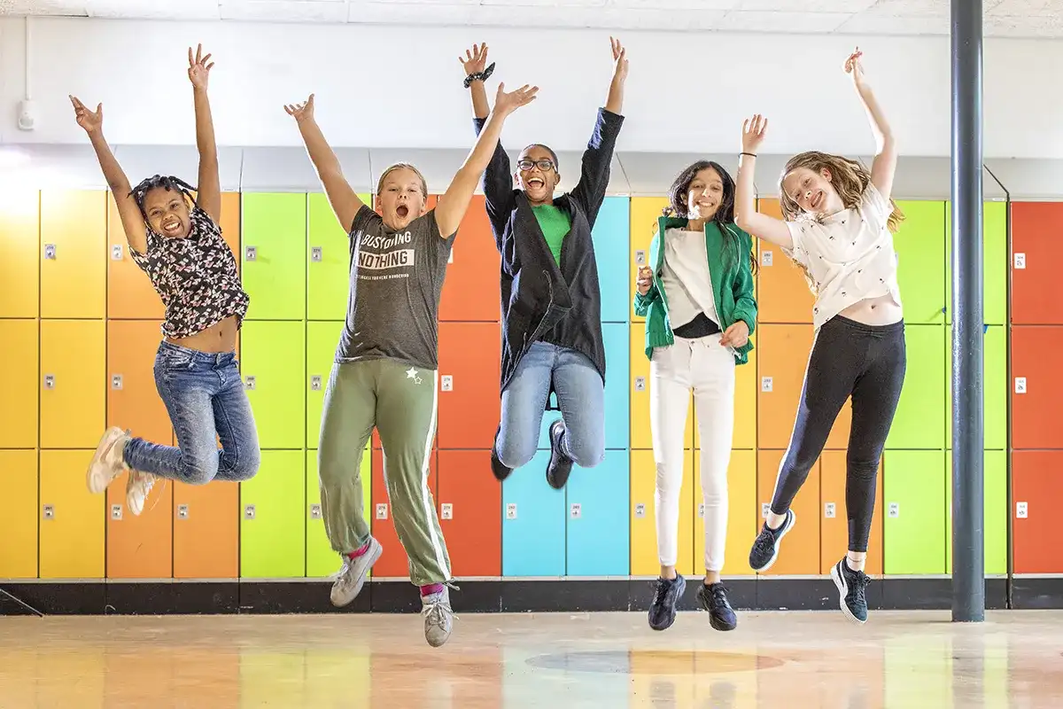 Vijf kinderen springen vrolijk in de lucht voor een muur van kleurrijke kluisjes. Ze dragen allemaal vrijetijdskleding en glimlachen breed, hun armen hoog geheven. De vloer is een glanzend binnenoppervlak en ze worden midden in de sprong vastgelegd, wat een gevoel van opwinding en geluk uitbeeldt.