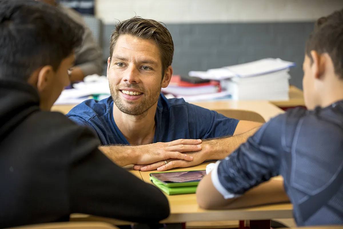 Een glimlachende man met kort bruin haar zit aan een tafel en is in gesprek met twee zittende personen die met de rug naar de camera staan. Op de tafel liggen diverse papieren en mappen, en op de achtergrond liggen nog meer stapels papieren. De setting lijkt op een klaslokaal of studieomgeving.