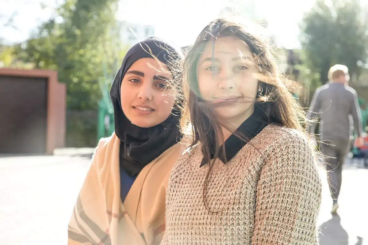 Twee vrouwen zitten buiten en lachen naar de camera. De vrouw links draagt een hijab en een beige, blauwe en rode sjaal. De vrouw rechts heeft lang, lichtbruin haar dat in de wind wappert en is gekleed in een beige trui. De achtergrond bestaat uit groen en een voorbijlopende persoon.
