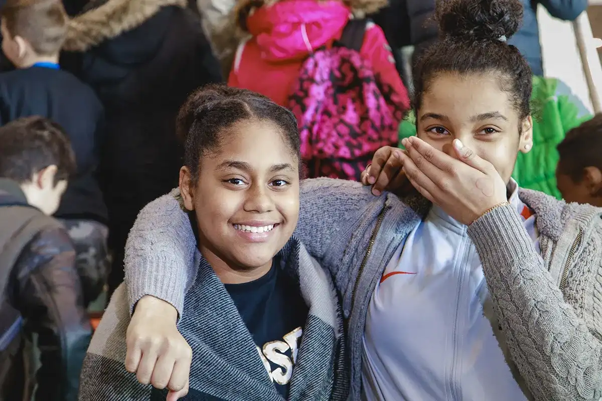 Twee kinderen staan glimlachend dicht bij elkaar. Het kind links draagt een grijs-zwart jasje over een zwart shirt. Het kind rechts, gekleed in een wit jasje met ritssluiting, bedekt met één hand zijn mond. Beiden hebben donker haar in knotjes. Wazige mensen en rugzakken zijn zichtbaar op de achtergrond.
