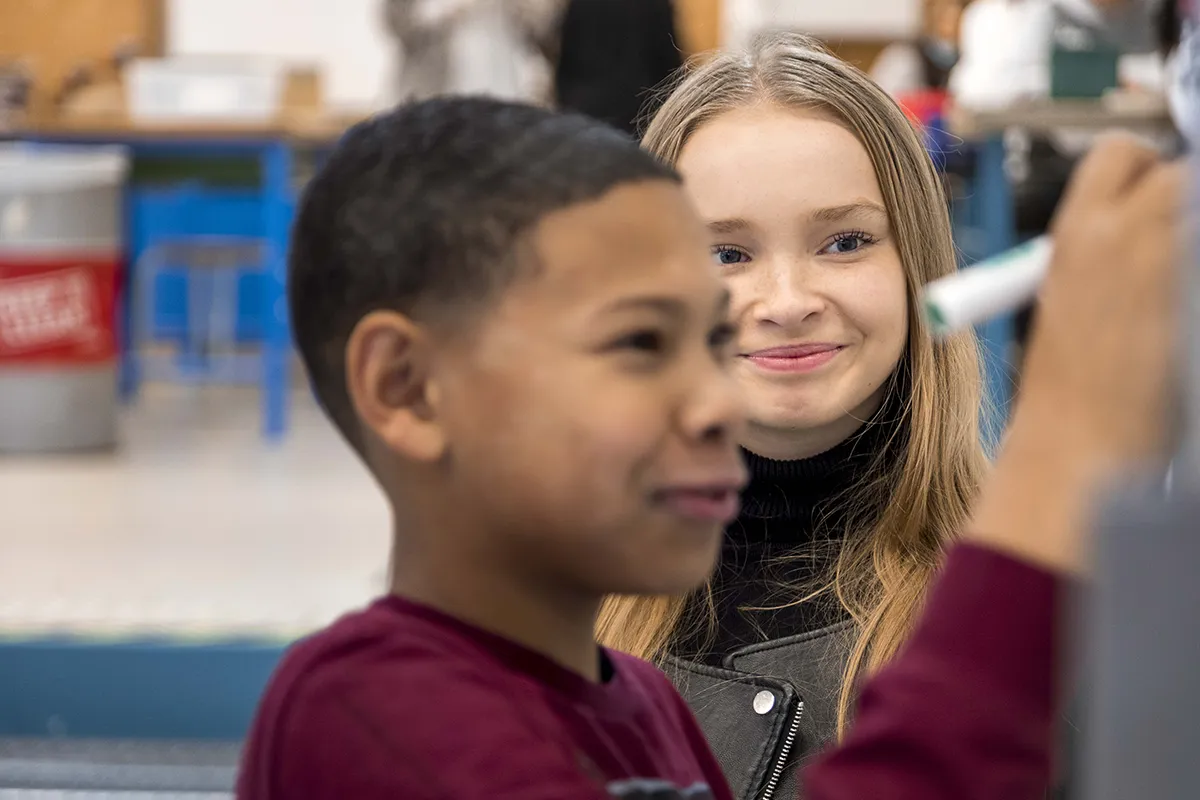 Een jonge jongen met kort zwart haar en een kastanjebruin shirt draagt, schrijft met een stift op een whiteboard. Achter hem staat een meisje met lang blond haar glimlachend naar hem te kijken. Ze bevinden zich in wat lijkt op een klaslokaal met een onscherpe achtergrond, inclusief bureaus en stoelen.