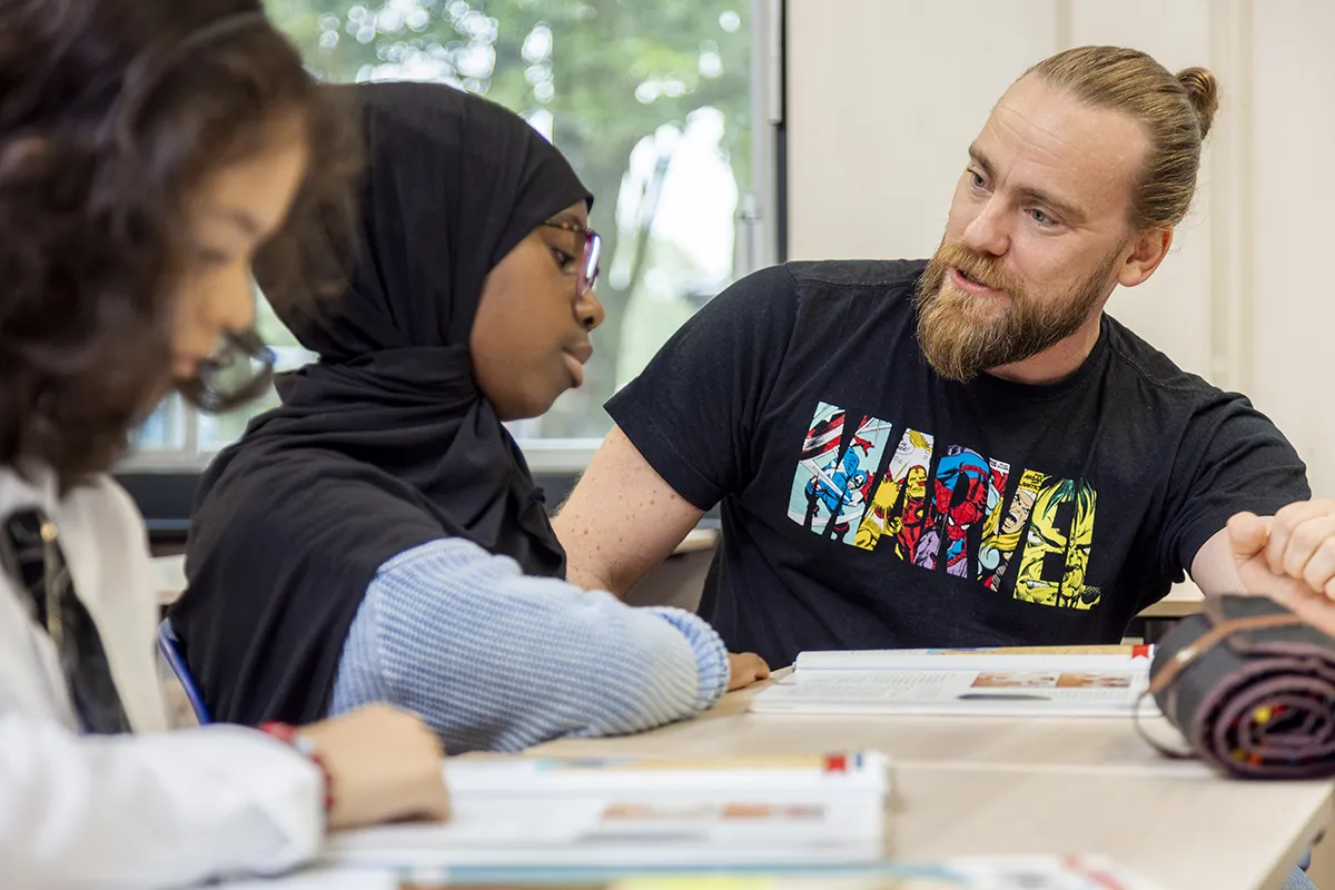 Een man met baard en samengebonden haar, gekleed in een Marvel-T-shirt, zit met twee meisjes aan een tafel. Eén meisje, gekleed in een hijab en een bril, praat met de man. Het andere meisje, in een wit overhemd en stropdas, is gefocust op haar werk. Boeken en schoolspullen liggen voor hen op tafel uitgespreid.