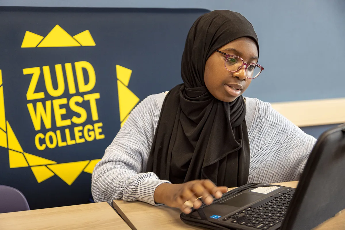 Een student met een zwarte hijab en bril zit voor een laptop te typen. Ze draagt een grijze trui en zit in een klaslokaal. Achter haar staat een donkerblauwe muur met het gele logo van 'Zuid West College'. Ze lijkt gefocust en betrokken bij haar werk.