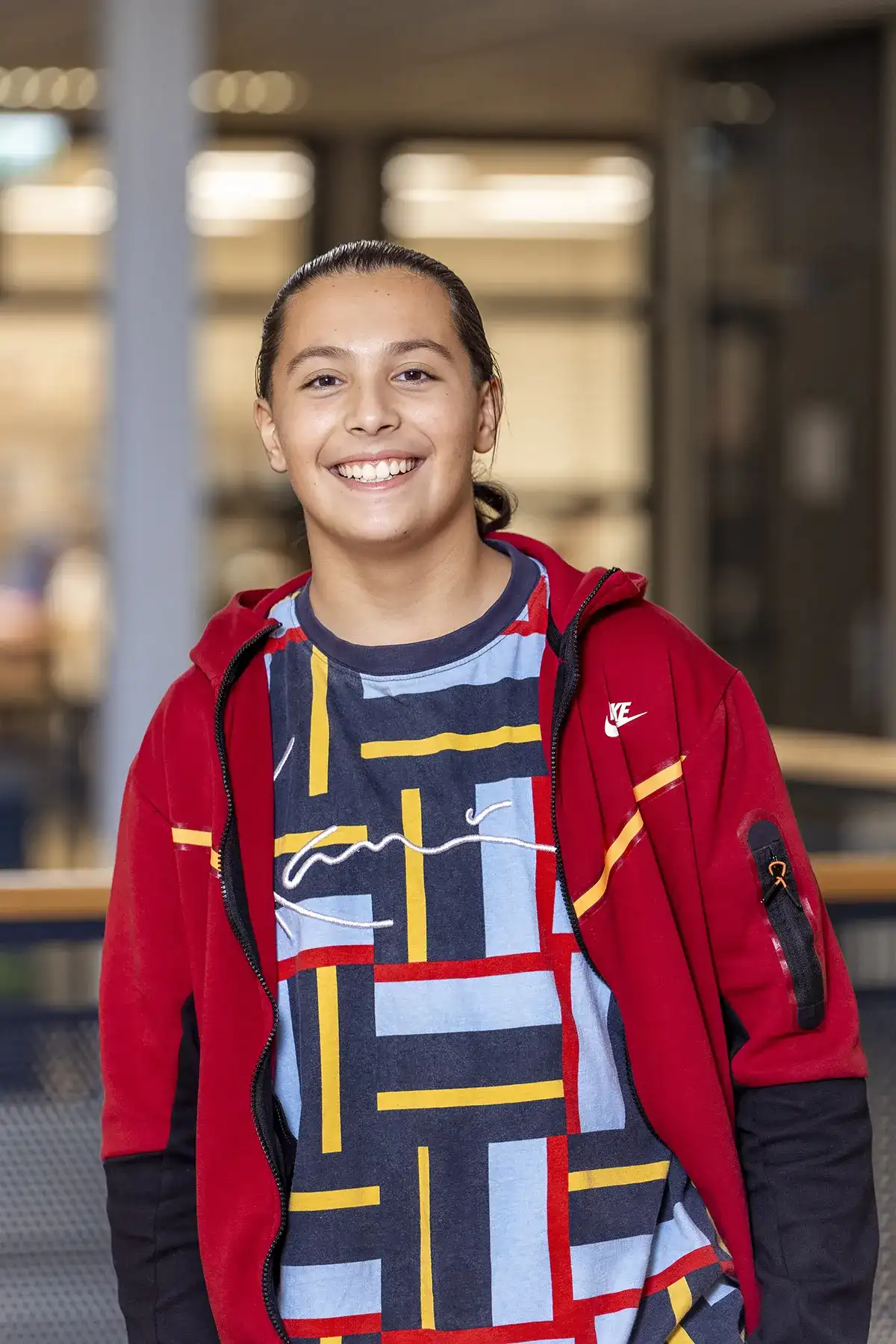 Een persoon met lang achterover gebonden haar lacht naar de camera. Ze dragen een rood jasje over een grafisch T-shirt met een veelkleurig geometrisch patroon van lijnen en rechthoeken. De achtergrond lijkt een binnenomgeving te zijn met vage details, waarschijnlijk een kantoor- of schoolomgeving.