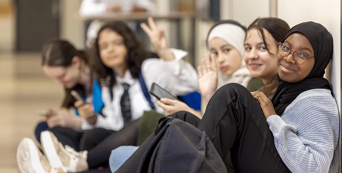 Een diverse groep van vijf jonge vrouwen zit op de grond in een gang. Sommigen zwaaien en gebaren vredestekens, terwijl anderen naar hun telefoons kijken. Ze zijn allemaal nonchalant gekleed, de een draagt een hijab en de ander een hoofddoek. De sfeer is ontspannen en vriendelijk.