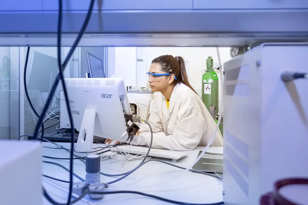 Een wetenschapper in een witte laboratoriumjas en veiligheidsbril concentreert zich aandachtig op een computerscherm op een laboratoriumwerkplek. Om haar heen zijn diverse wetenschappelijke apparatuur, kabels en gasflessen zichtbaar. De setting suggereert een hightech onderzoeksomgeving.