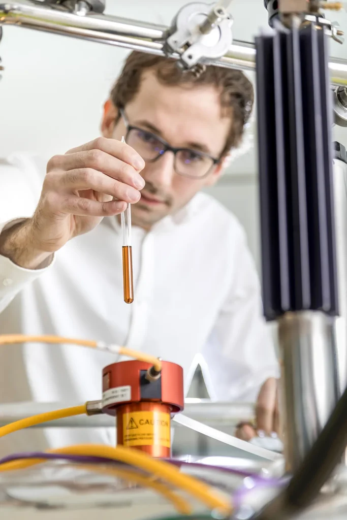 Een wetenschapper met een bril en een witte laboratoriumjas houdt zorgvuldig een pipet met daarin een oranje vloeistof vast. De laboratoriumapparatuur omvat verschillende buisjes en containers, waarvan er één is voorzien van een waarschuwingsbord. Op de achtergrond zijn extra wetenschappelijke apparaten en metalen constructies te zien.