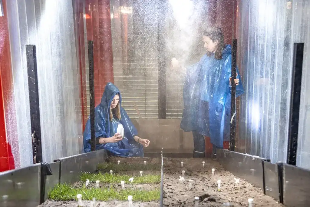 Twee personen, gekleed in blauwe regenponcho's, bevinden zich in een transparant bouwwerk waar watermist rond spuit. Eén persoon hurkt en houdt een klein wit voorwerp vast, terwijl de ander vlakbij staat met de deur op een kier. De grond binnenin heeft plekken met gras en kale grond met witte markeringen.