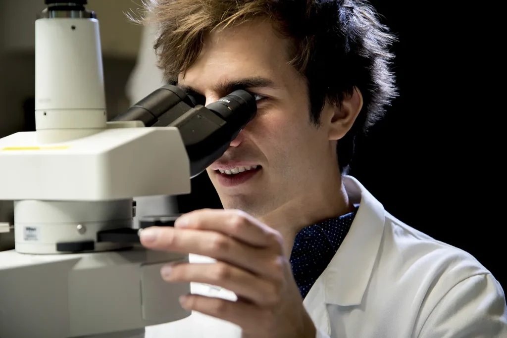 Een persoon met halflang bruin haar kijkt door een microscoop in een laboratoriumomgeving. Ze dragen een witte laboratoriumjas en stellen scherp op het oculair van de microscoop terwijl ze met hun rechterhand de focusknop verstellen. De achtergrond is zwak verlicht, waardoor de microscoop en de persoon worden benadrukt.