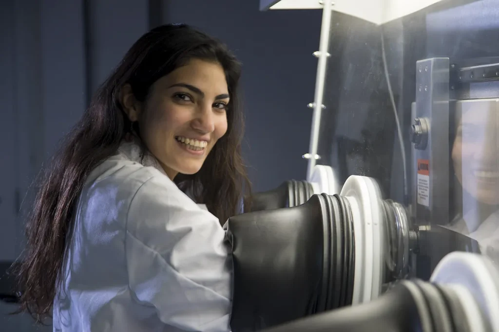 Een vrouw met lang donker haar glimlacht terwijl ze in een laboratorium werkt. Ze draagt een witte laboratoriumjas en gebruikt handschoenpoorten in een afgesloten insluitingskamer. De verlichting rondom haar werkruimte is helder en op de achtergrond zijn diverse wetenschappelijke apparatuur zichtbaar.