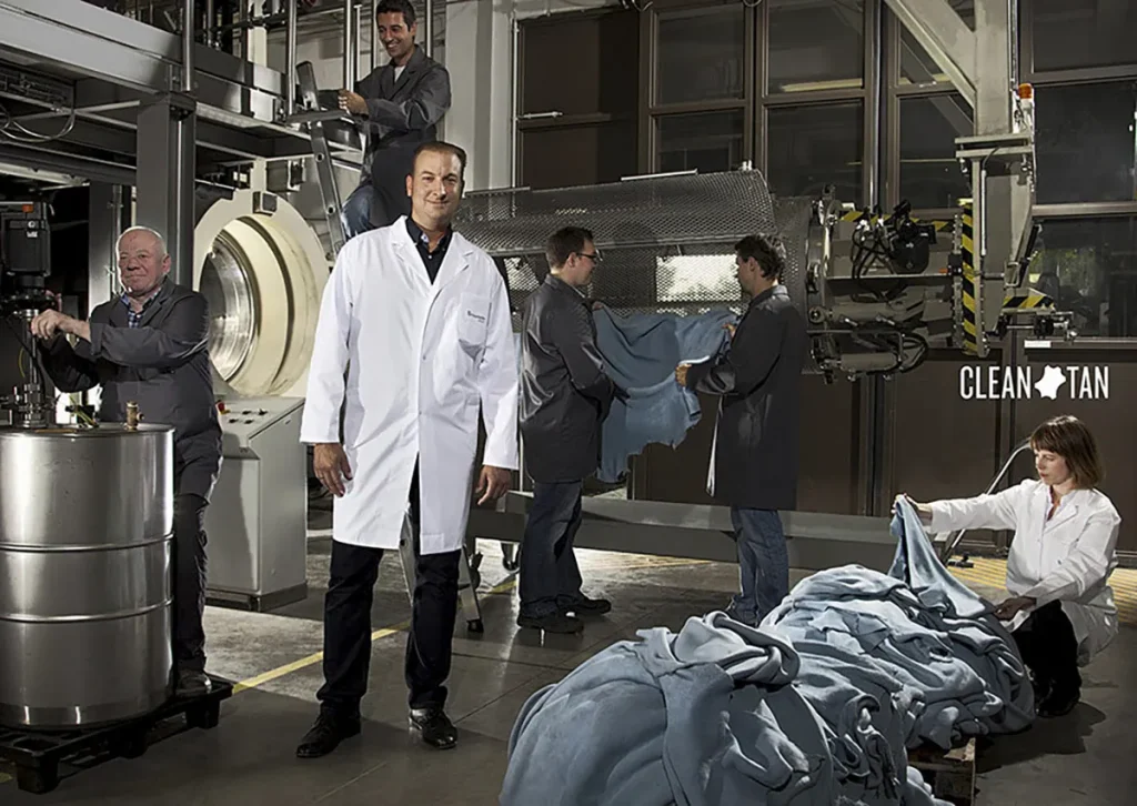 Een groep van vijf personen werkt in een industriële omgeving. Eén persoon staat vooraan, gekleed in een witte laboratoriumjas en glimlachend. Twee mensen op de achtergrond bedienen machines, terwijl twee anderen een grote stapel blauwe stof beheren met het opschrift 'Clean Tan'. De setting lijkt een textiel- of stoffenverwerkingsfaciliteit te zijn.