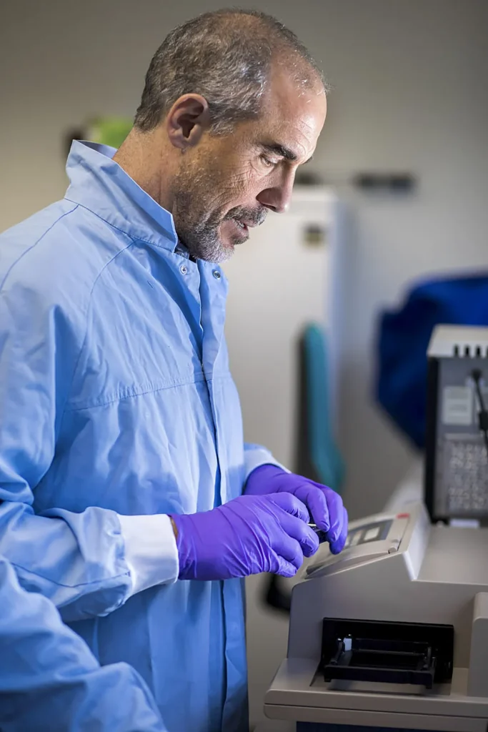Een persoon met kort grijs haar en een baard, gekleed in een lichtblauwe laboratoriumjas en paarse handschoenen, bedient een laboratoriummachine. Ze drukken met gerichte aandacht op knoppen op een bedieningspaneel. De achtergrond bestaat uit wazige laboratoriumapparatuur, wat een professionele laboratoriumomgeving suggereert.