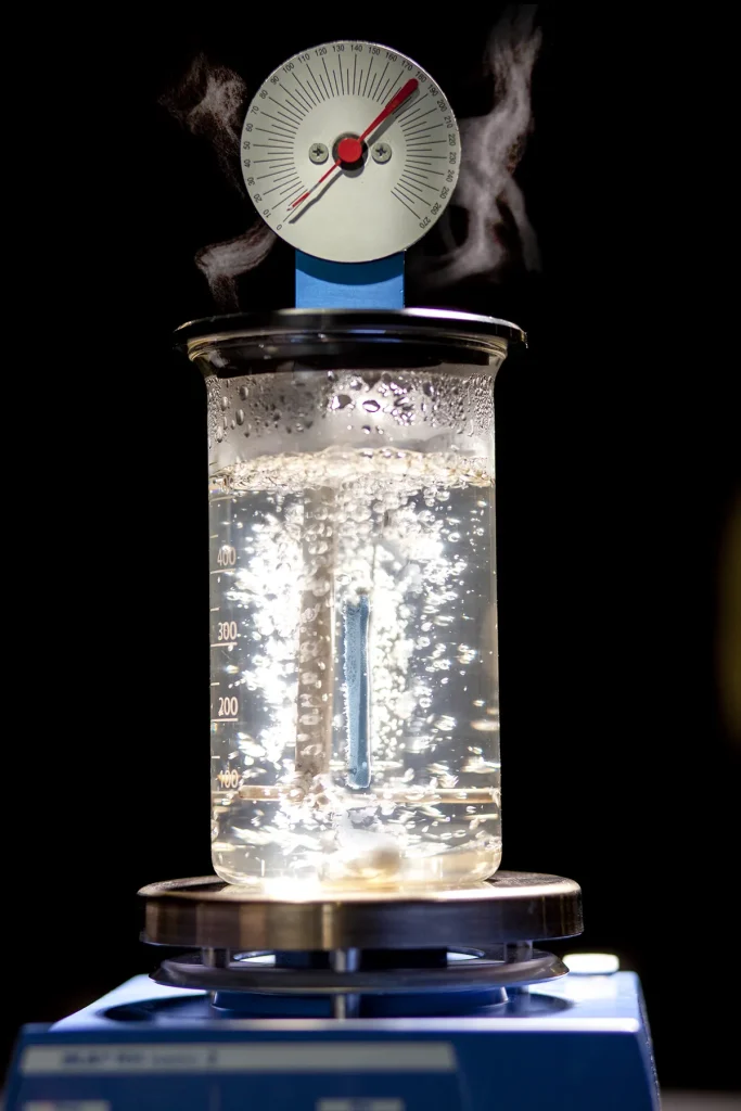 Een glazen beker gevuld met kokend water staat op een blauwe laboratoriumkookplaat. Stoom stijgt op en bellen stijgen krachtig op in de beker. Boven de beker meet een thermometer met een rode naald de temperatuur. De achtergrond is zwart, wat de laboratoriumopstelling benadrukt.