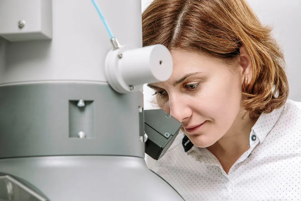 Een vrouw met kort bruin haar in een wit overhemd met lange mouwen en stippen kijkt door de lens van een groot, grijs wetenschappelijk instrument, waarschijnlijk een microscoop. Haar gezicht toont concentratie en de machine bevat verschillende knoppen en opzetstukken. De achtergrond is duidelijk en onscherp.