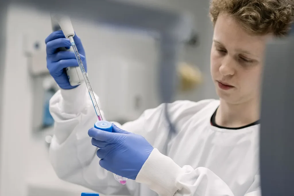 Een persoon die een witte laboratoriumjas en blauwe handschoenen draagt, gebruikt een pipet om een roze vloeistof over te brengen in een kleine, doorzichtige container in een laboratoriumomgeving. Ze lijken gefocust op hun taak, terwijl op de onscherpe achtergrond verschillende laboratoriumapparatuur zichtbaar is.