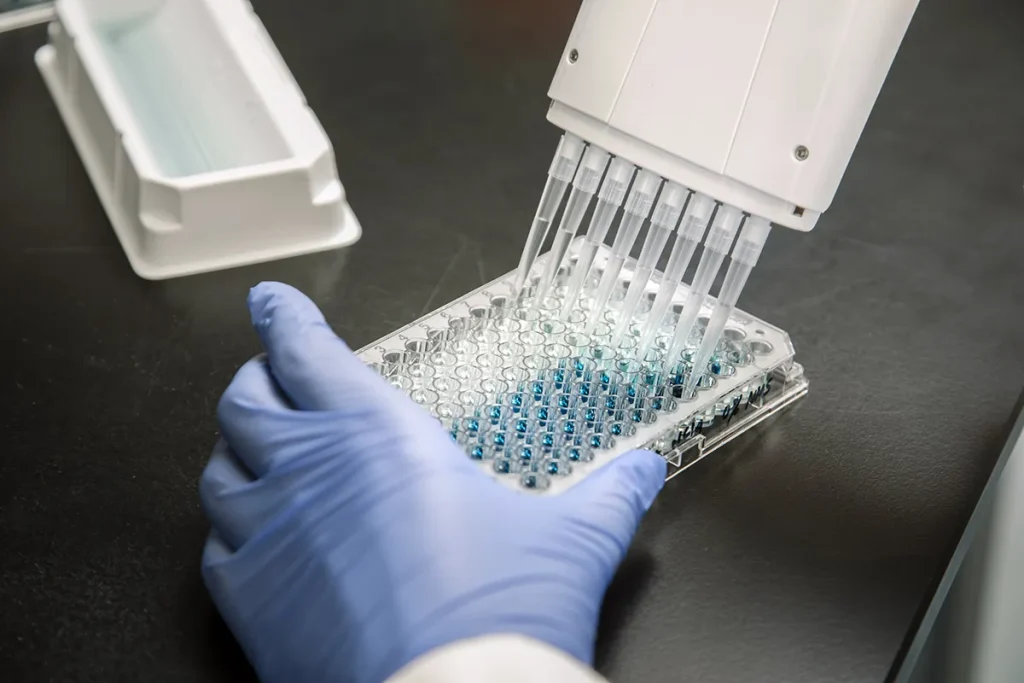 Een gehandschoende hand houdt een plaat met 96 putjes vast met blauwe vloeistof in elk putje, geplaatst onder een meerkanaalspipetdispenser. De pipet verdeelt vloeistof in de putjes. De opstelling wordt op een donkere laboratoriumbank geplaatst met een witte container op de achtergrond.