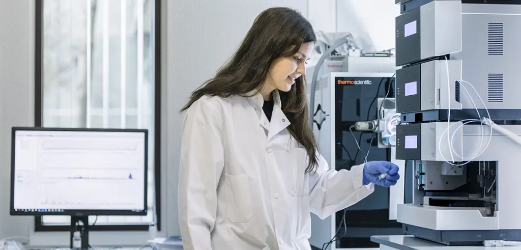 Een wetenschapper in een laboratoriumjas en blauwe handschoenen bedient een krachtige vloeistofchromatografiemachine (HPLC). Ze staat naast een computer waarop analyseresultaten worden weergegeven en glimlacht terwijl ze de apparatuur aanpast. De laboratoriumomgeving omvat verschillende wetenschappelijke instrumenten en een raam op de achtergrond.