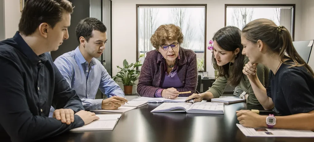 In een kantoor zitten vijf mensen rond een tafel. Vier jongvolwassenen, drie mannen en één vrouw, luisteren aandachtig naar een oudere vrouw met krullend haar die spreekt en gebaart naar een open boek. Papieren en notitieboekjes liggen verspreid op tafel en op de achtergrond zijn planten zichtbaar.