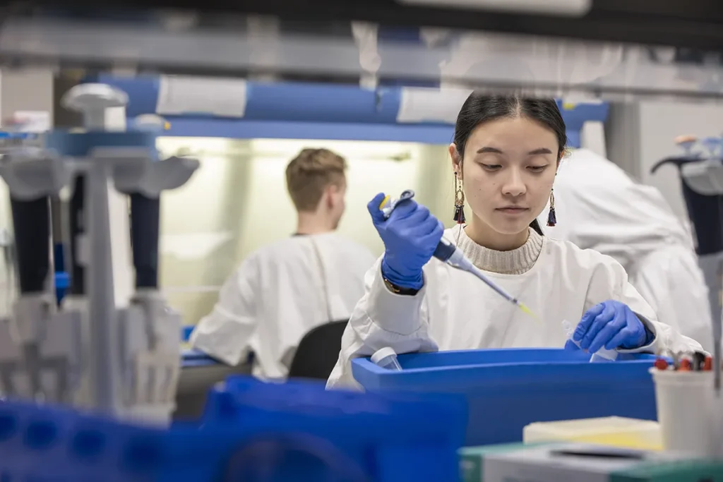 Een wetenschapper in een witte laboratoriumjas en blauwe handschoenen gebruikt een pipet in een onderzoekslaboratorium. Ze is gefocust op haar taak. Op de achtergrond werkt een andere wetenschapper op een laboratoriumbank, gedeeltelijk onscherp. Het laboratorium is uitgerust met diverse wetenschappelijke instrumenten en benodigdheden.