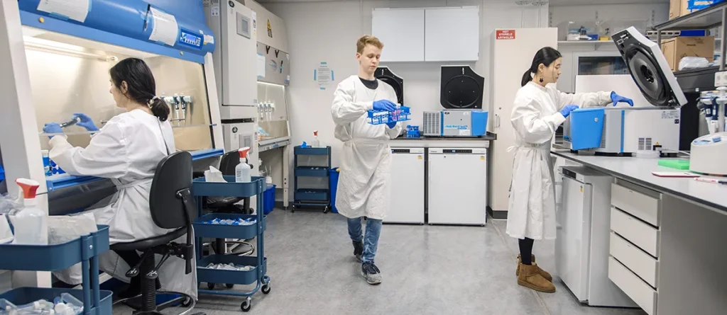 Drie wetenschappers werken in een modern laboratorium. Twee vrouwen voeren taken uit op aparte laboratoriumstations, terwijl een man in het midden loopt met blauwe dozen met laboratoriummateriaal. Iedereen is gekleed in laboratoriumjassen, handschoenen en een veiligheidsbril. Het laboratorium beschikt over geavanceerde apparatuur en opbergkasten.