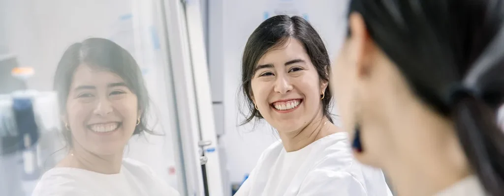 Een vrouw met donker haar, gekleed in een witte jas, glimlacht terwijl ze naar haar spiegelbeeld kijkt op een glazen oppervlak in een laboratoriumomgeving. Ze staat in profiel, maar haar gezicht is volledig zichtbaar in de weerspiegeling. Laboratoriumapparatuur en documenten zijn onduidelijk zichtbaar op de achtergrond.