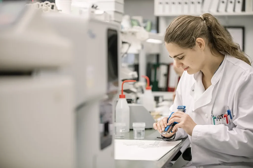 Een wetenschapper in een witte laboratoriumjas is gefocust op haar werk in een laboratorium. Ze houdt een container met een blauwe dop vast en lijkt de inhoud ervan te onderzoeken of te testen. Het laboratorium is uitgerust met verschillende instrumenten, benodigdheden en documenten. De omgeving is georganiseerd en goed verlicht.