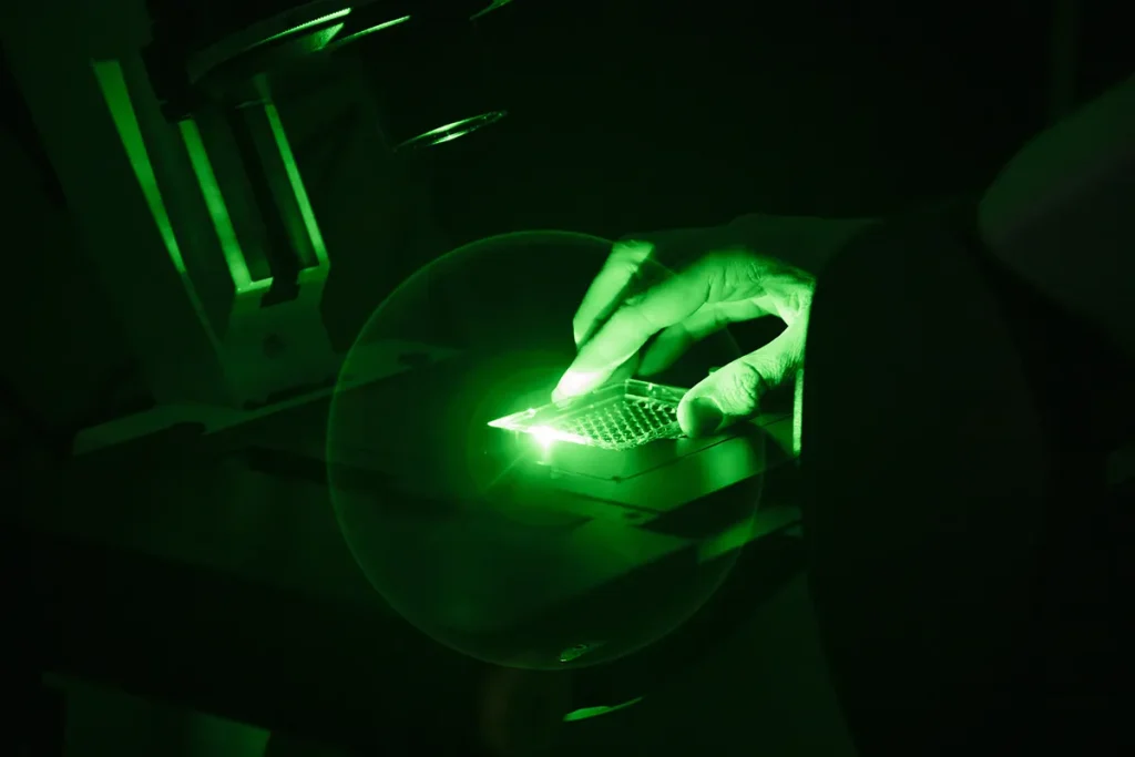 Een close-up van een hand onder groen licht, die werkt met een klein elektronisch onderdeel of een printplaat. De setting lijkt een verduisterd laboratorium te zijn met gespecialiseerde apparatuur, mogelijk een microscoop, rond het gebied waar het werk wordt uitgevoerd.