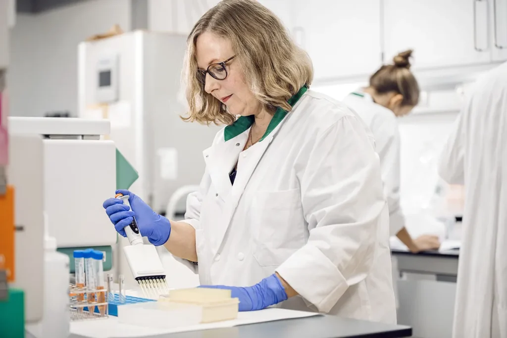 Een vrouwelijke wetenschapper met schouderlang haar en een bril, gekleed in een witte laboratoriumjas en blauwe handschoenen, gebruikt een pipet in een laboratorium. Reageerbuisjes en apparatuur staan voor haar op de werkbank. Op de achtergrond is nog een laboratoriummedewerker zichtbaar, eveneens gekleed in een laboratoriumjas.