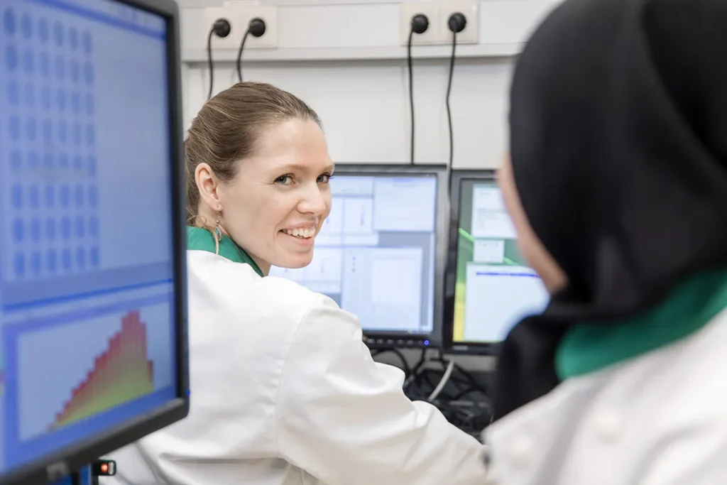 Een zittende vrouw in een witte laboratoriumjas lacht en kijkt naar een collega. Ze wordt geconfronteerd met een andere persoon die een soortgelijke jas en een zwarte hoofddoek draagt. Ze werken aan bureaus met computers die gegevens en grafieken weergeven. De omgeving lijkt een laboratorium- of kantooromgeving te zijn.