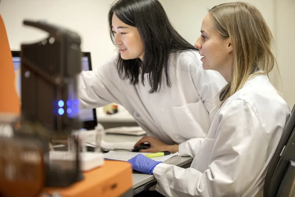 Twee wetenschappers werken in een laboratorium. De ene vrouw wijst naar een computerscherm terwijl de ander ernaar kijkt, beiden gekleed in witte laboratoriumjassen. Op de voorgrond staat laboratoriumapparatuur en beide lijken gefocust op hun taak. De één heeft lang donker haar en de ander heeft lang blond haar.