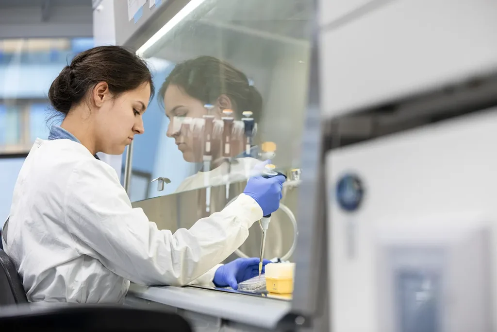 Een onderzoeker in een laboratoriumjas en blauwe handschoenen werkt bij een bioveiligheidskabinet in een laboratorium. Ze gebruikt een pipet om vloeistof in een container over te brengen. De kast is voorzien van reflecterend glas en op de voor- en achtergrond is diverse laboratoriumapparatuur zichtbaar.