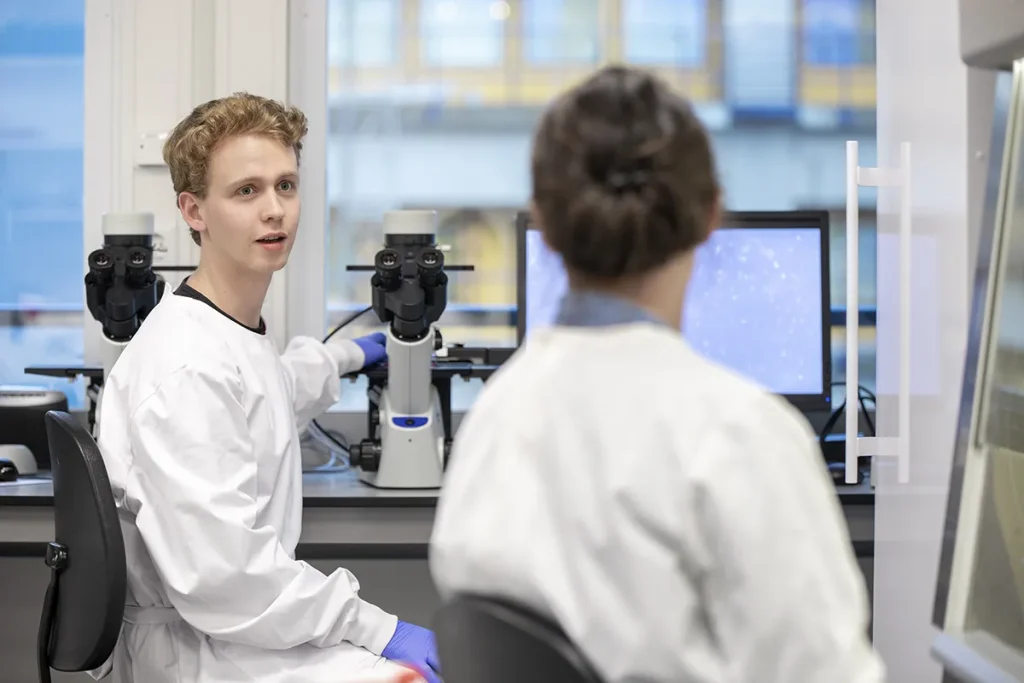 Twee wetenschappers in witte laboratoriumjassen werken in een laboratorium. Eén, vanaf de achterkant gezien, zit voor een computerscherm met blauw-witte afbeeldingen. De ander, een jonge man met krullend haar, zit achter een microscoop en kijkt terwijl hij spreekt naar zijn collega.