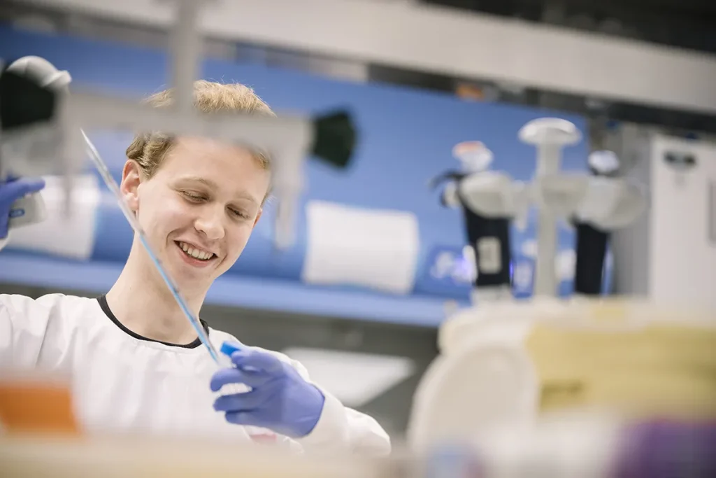 Een persoon in een laboratoriumjas en blauwe handschoenen glimlacht terwijl hij in een laboratorium werkt. Ze houden een pipet vast en lijken een experiment uit te voeren. Op de onscherpe achtergrond zijn diverse laboratoriumapparatuur, waaronder pipetten en containers, zichtbaar. De setting is gericht op wetenschappelijk onderzoek.