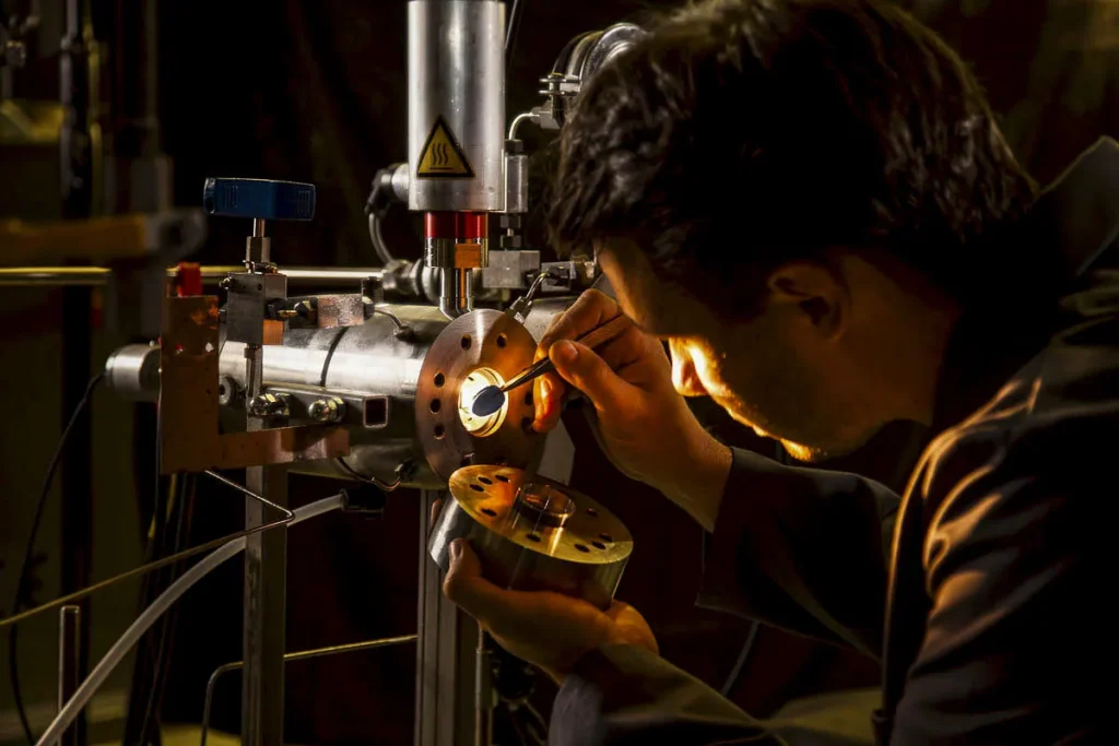 Een man werkt aandachtig met precieze instrumenten aan een machine in een slecht verlichte werkplaats. Met een klein gereedschap past hij zorgvuldig een metalen onderdeel aan. De machinerie is complex, met verschillende pijpen, bouten en een waarschuwingslabel. De scène wordt verlicht door gerichte verlichting.