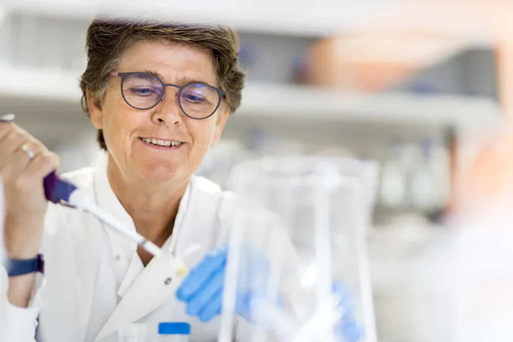 Een persoon met kort haar en een bril, gekleed in een witte laboratoriumjas, glimlacht terwijl hij een pipet hanteert in een laboratorium. De achtergrond is wazig, maar toont verschillende laboratoriumapparatuur, wat duidt op een wetenschappelijke setting.