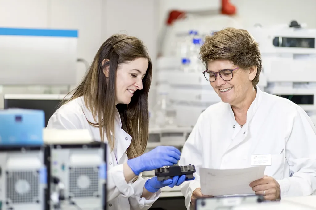 In een laboratorium werken twee vrouwen in witte laboratoriumjassen. De vrouw aan de linkerkant, met lang donker haar, draagt blauwe handschoenen en houdt een laboratoriuminstrument vast, glimlachend en laat het aan haar collega zien. De oudere vrouw aan de rechterkant, met kort bruin haar en een bril, houdt een document vast en glimlacht ook. Op de achtergrond is laboratoriumapparatuur zichtbaar.