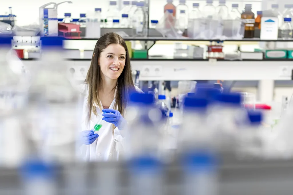 Een lachende vrouw in een witte laboratoriumjas en blauwe handschoenen staat in een laboratorium en houdt een groene vloeistof in een reageerbuis. Planken achter haar zijn gevuld met flessen en laboratoriumapparatuur. Wazige voorgrondelementen, waaronder meer flessen, omlijsten de heldere en overzichtelijke laboratoriumomgeving.