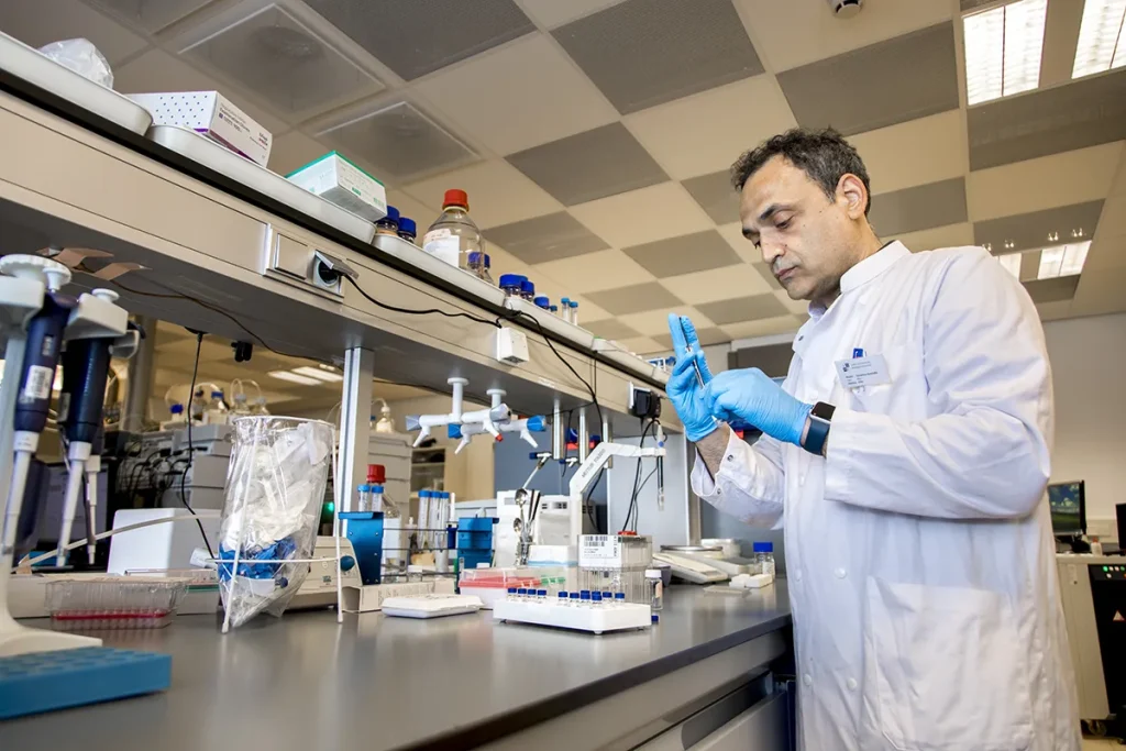 Een wetenschapper in een witte laboratoriumjas en blauwe handschoenen werkt in een modern laboratorium. Ze hanteren een pipet en kijken naar een klein bakje, waarschijnlijk bezig met een experiment. De laboratoriumbank staat vol met wetenschappelijke apparatuur, waaronder pipetrekken, reageerbuisjes en containers.
