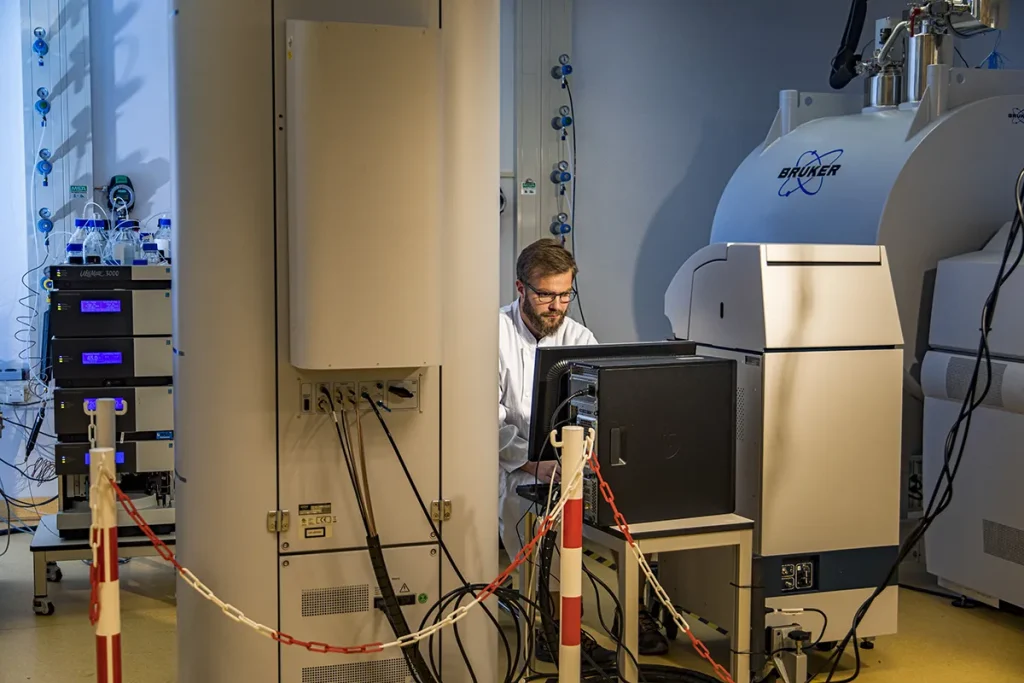 Een wetenschapper in een witte laboratoriumjas en bril werkt op een computer in een laboratorium. Om hem heen bevindt zich geavanceerde wetenschappelijke apparatuur, waaronder een grote Bruker-machine, elektrische kasten en andere laboratoriumapparatuur. Voor de veiligheid is het gebied afgezet met rode en witte kettingen.