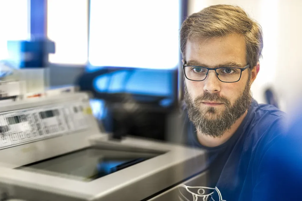 Een man met baard en bril kijkt aandachtig naar een machine, mogelijk voor wetenschappelijk of technisch werk. De achtergrond is wazig en de grote ramen laten fel licht binnen. Hij draagt een donker T-shirt en de omgeving lijkt een laboratorium of werkruimte met diverse apparatuur.