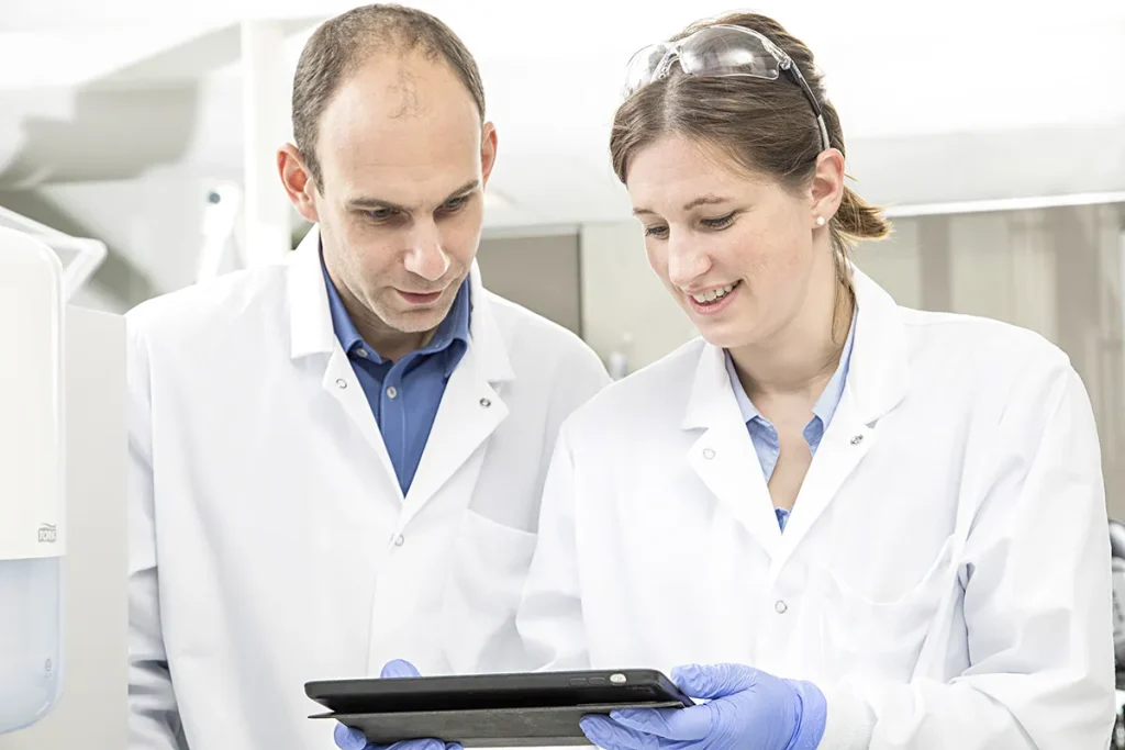 Twee wetenschappers in witte laboratoriumjassen zijn in een laboratorium. De man aan de linkerkant, kaal en gekleed in een blauw shirt, kijkt naar een tablet die wordt vastgehouden door de vrouw aan de rechterkant, die bruin haar heeft en een veiligheidsbril, een lichtblauw shirt en blauwe handschoenen draagt. Beiden glimlachen en zijn gefocust op de tablet.