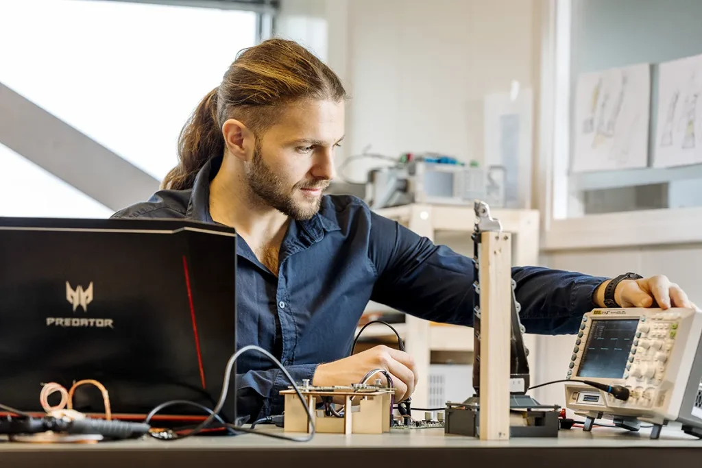 Een man met een paardenstaart, gekleed in een donkerblauw shirt, zit aan een tafel en werkt met elektronische apparatuur. Hij past een oscilloscoop aan terwijl hij omringd wordt door verschillende elektronische componenten en een laptop met het logo "Predator" zichtbaar. De achtergrond toont een heldere, moderne laboratoriumomgeving.